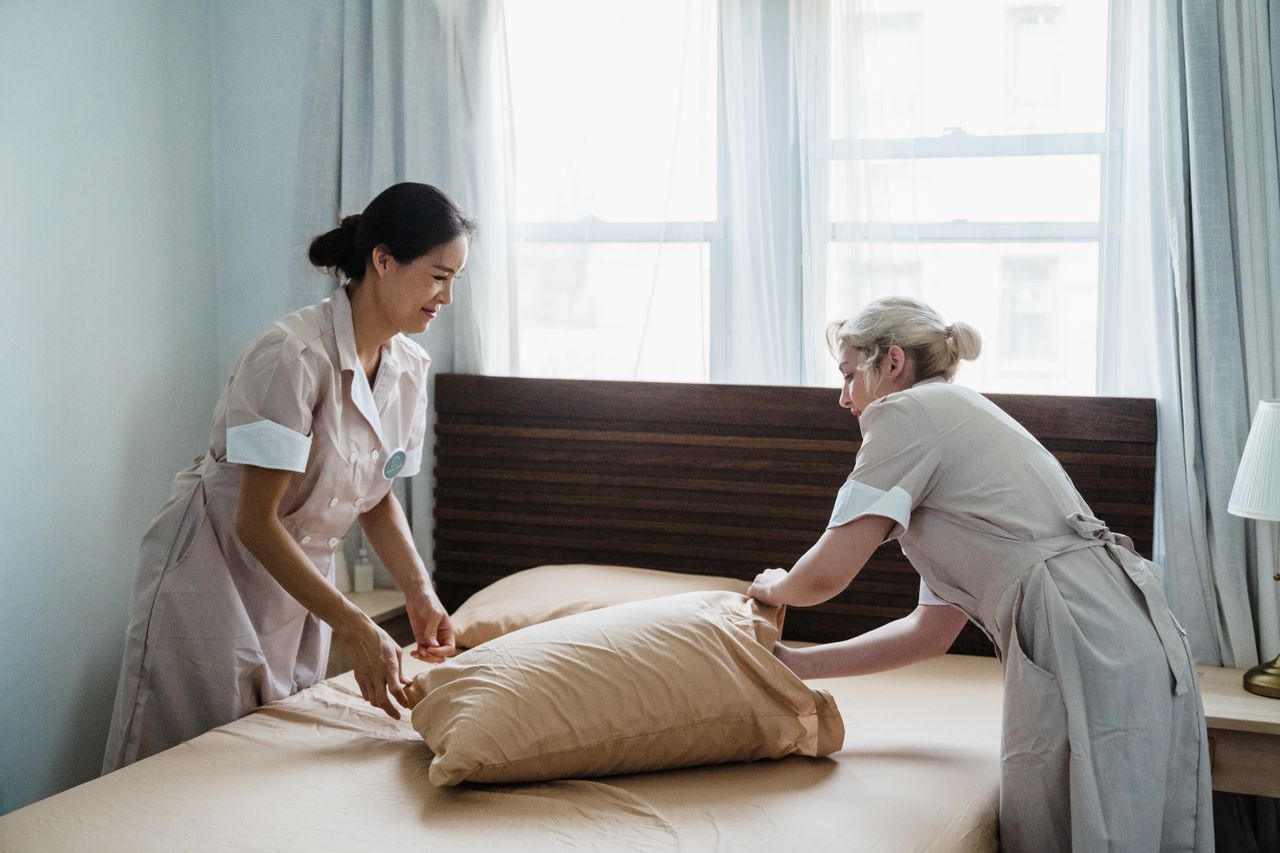 digital Two housekeepers in uniform collaborate while making a bed in a bright, peaceful bedroom.