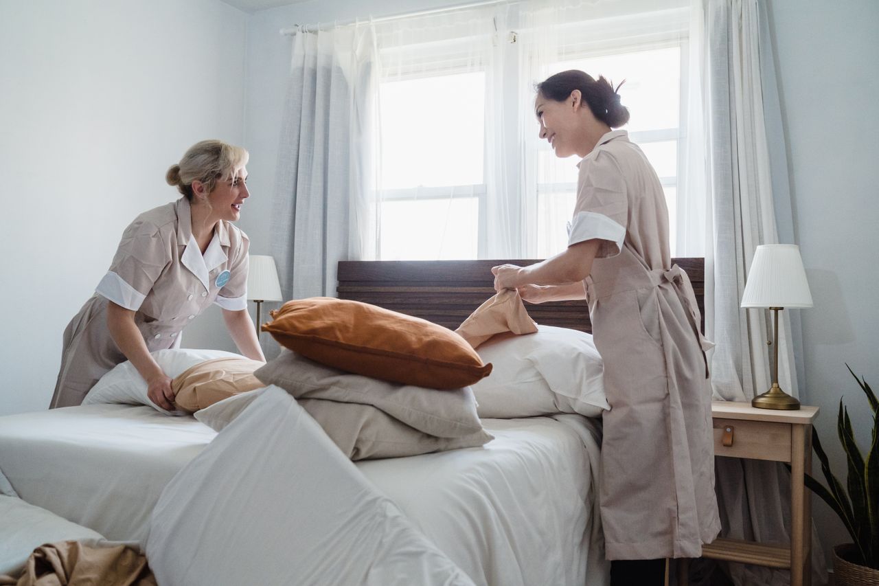 why-choose-us Two female housekeepers making a bed together in a well-lit room with tidy decor, showcasing teamwork.