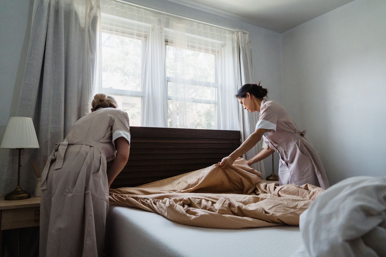 digital-01 Two housekeepers making a bed in a sunlit bedroom, showcasing cleanliness and attention to detail.