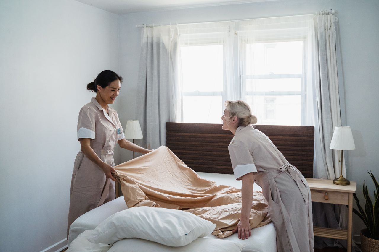 Services-01 Two housekeepers in uniform make a bed in a well-lit bedroom, showcasing efficient housekeeping service.