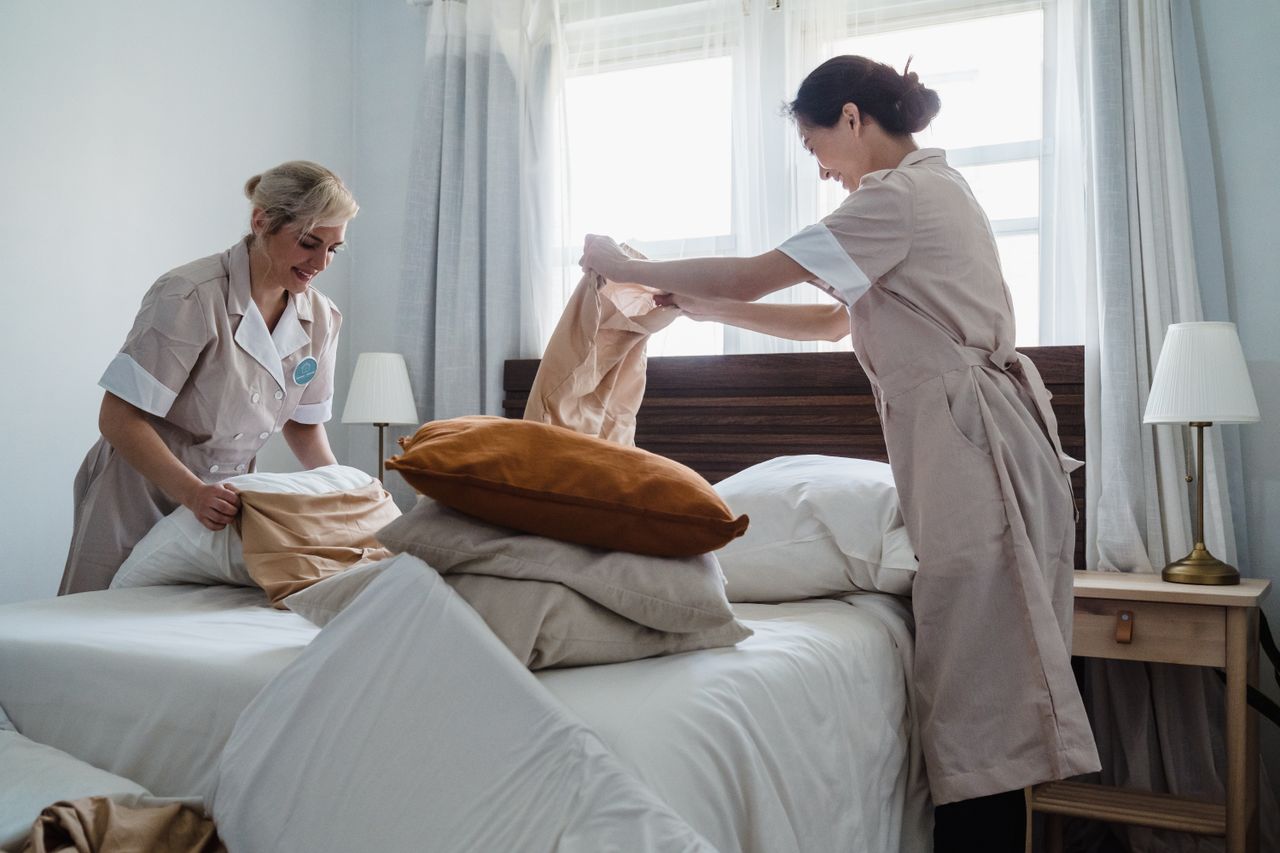Services-02 Two housekeepers in uniform fixing a hotel room bed with fresh linen and pillows.